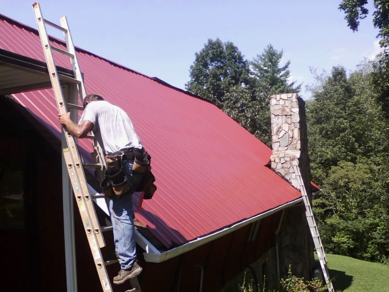 Roofer on a ladder beside a red metal roof and stone chimney — Soffit & Fascia Repair by Roofing Brothers in Rapid City